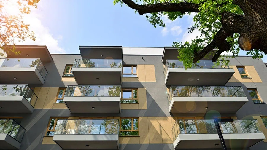 Moderne Mehrfamilienhausfassade mit großen Balkonen, viel Glas und warmem Sonnenlicht, während alte Bäume Schatten auf die oberen Etagen werfen.