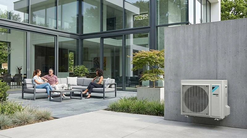 Vor einer modernen Glasfassade sitzt eine kleine Gruppe auf einer Terrasse, während rechts ein Daikin Außengerät an der Betonwand montiert ist.