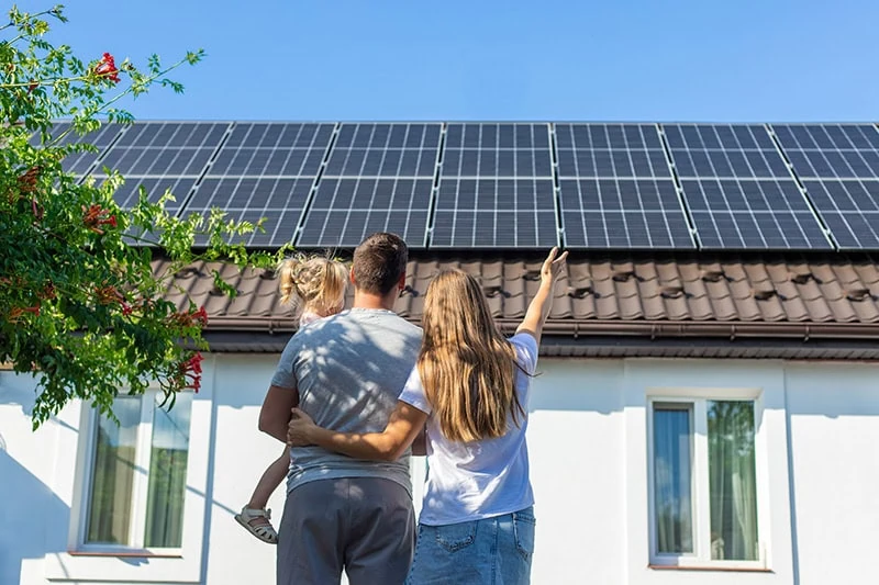 Eine Familie steht vor einem Haus mit Solarmodulen auf dem Dach, während die Frau auf die Anlage zeigt und der klare Himmel darüber leuchtet.