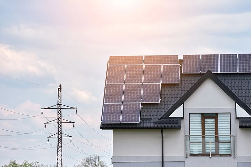 Auf einem modernen Hausdach liegen mehrere Solarmodule, während links ein Strommast unter einem hellen, leicht bewölkten Himmel steht.