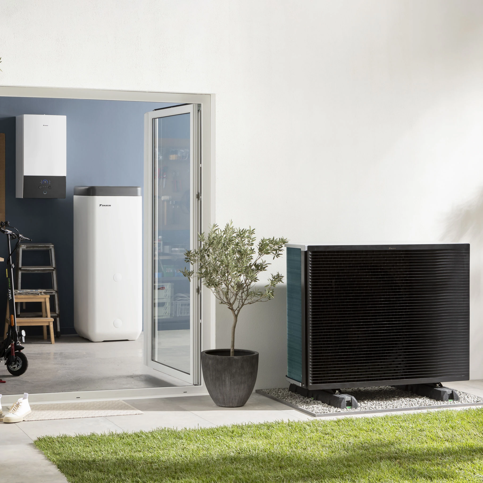 Vor einer weißen Hauswand stehen eine Daikin Wärmepumpe, ein Speicher und ein Außengerät neben Glas­tür, Pflanze und gepflegtem Rasen im Garten.