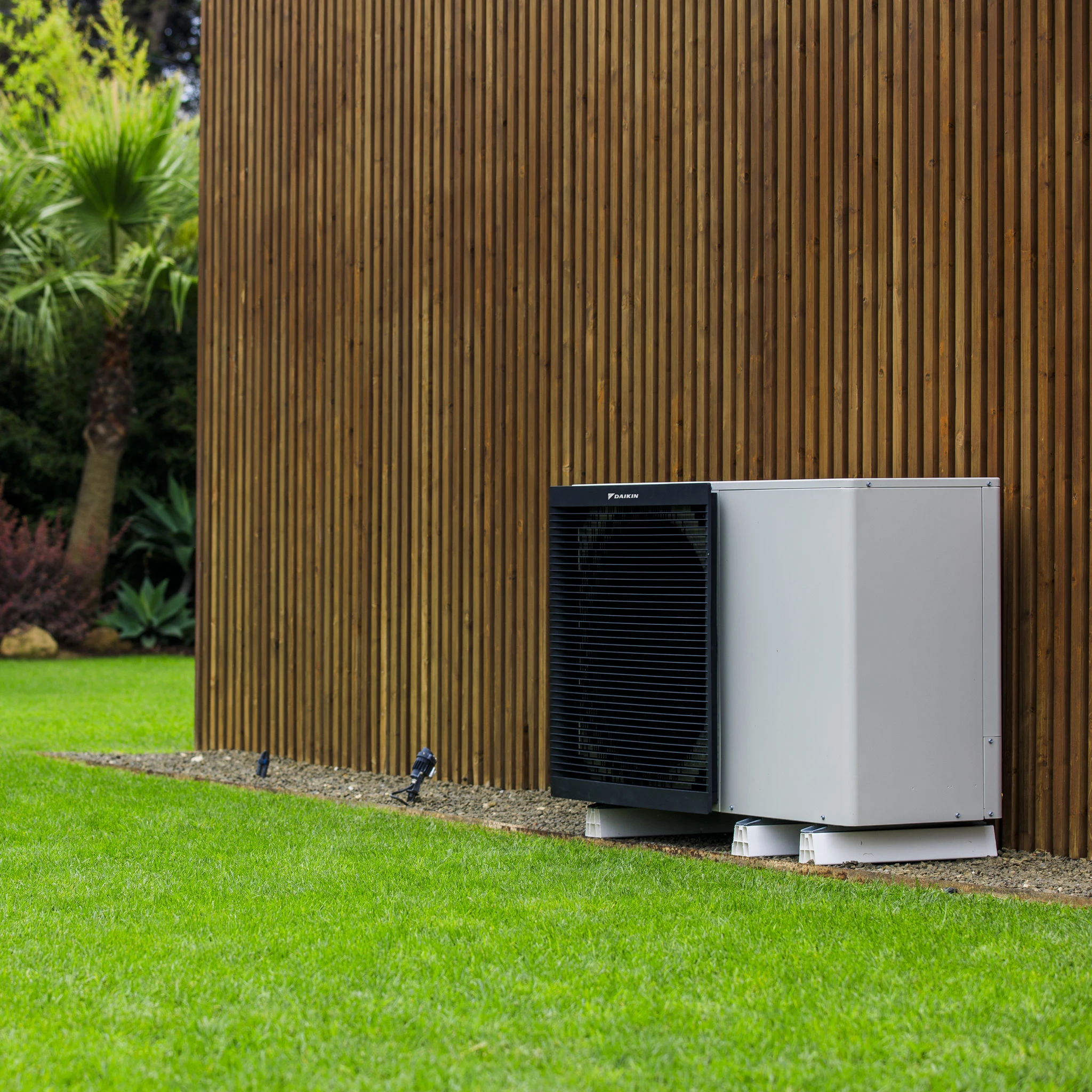 Eine Daikin Wärmepumpe steht vor einer Holzfassade im Garten, während grüner Rasen und tropische Pflanzen die moderne Außenanlage umgeben.